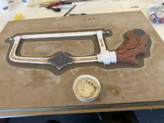 The bone saw frame part-painted on the bench beside a pot of weathering medium.