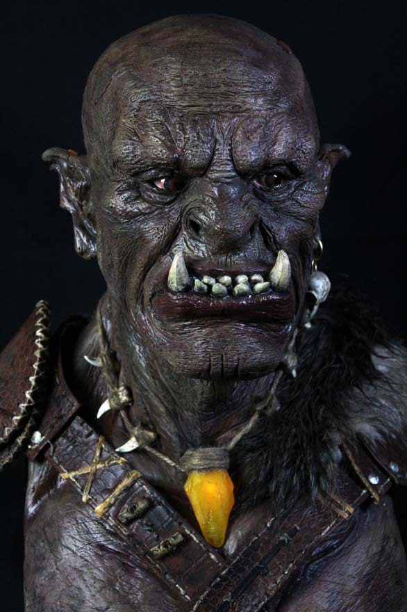 Front view of the painted orc bust showing tusks, layered skin colour and a brass pendant.